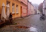 Brukowany plac w centrum małego miasta z pastelowymi kamienicami, ławką i starą pompą